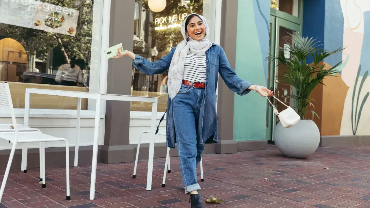 Fr&ouml;hliche Frau mit Hijab, die l&auml;ssig Boyfriend Jeans kombiniert. Sie tr&auml;gt eine Jeansjacke, gestreiftes Shirt, roten G&uuml;rtel und eine wei&szlig;e Tasche.