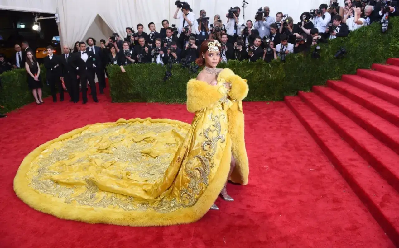 Rihanna in gelbem Kleid mit Schleppe und Pelzmantel auf rotem Teppich, umgeben von Fotografen. Ein echter Hingucker, wie ein gelber law roach.