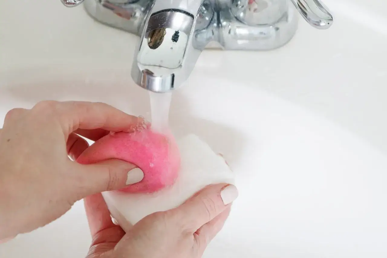 H&auml;nde reinigen einen pinken Make-up-Schwamm mit Seife unter flie&szlig;endem Wasser in einem wei&szlig;en Waschbecken.