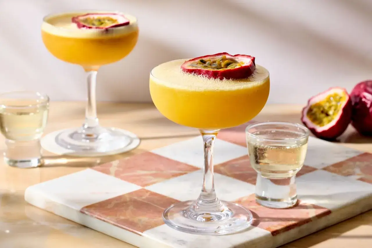 Zwei leuchtend gelbe Cocktails, garniert mit Passionsfrucht, bereit f&uuml;r das Pornstar Martini Rezept. Daneben kleine Gl&auml;ser mit Champagner.