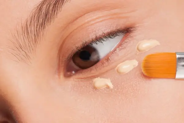 Nahaufnahme eines Auges, auf das mit einem Pinsel Concealer aufgetragen wird. Der Concealer setzt sich ab und bildet kleine Tupfen.