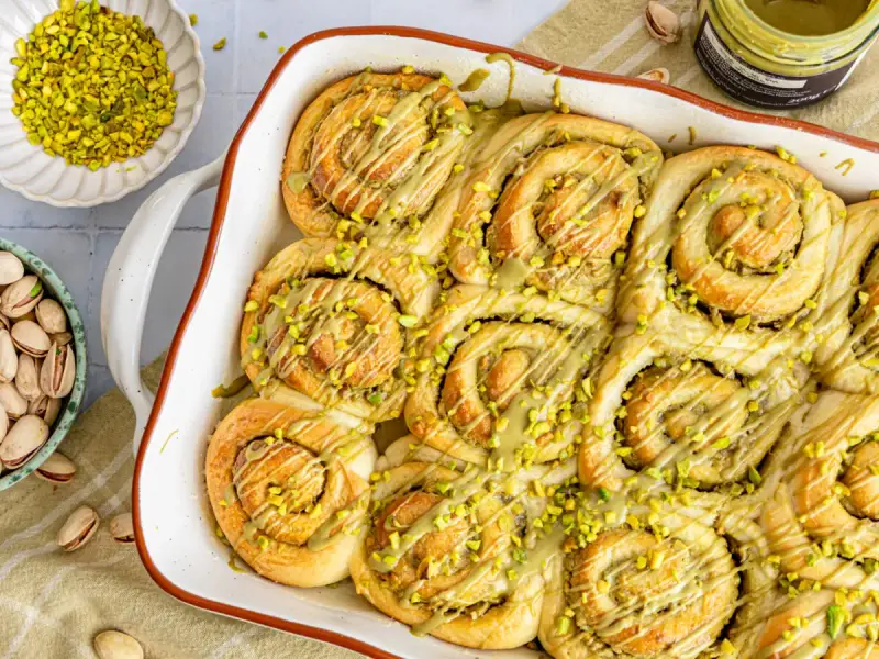 Frisch gebackene Pistazienschnecken, beträufelt mit Pistaziencreme und bestreut mit gehackten Pistazien. Daneben Schalen mit ganzen und gehackten Pistazien.