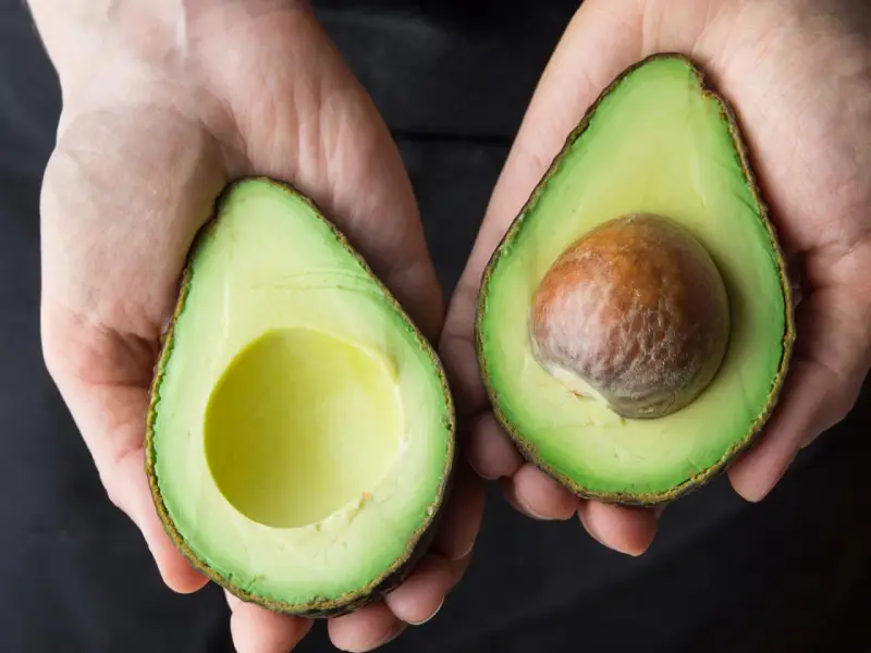 Hände halten zwei Avocadehälften. Eine Avocado mit Kern, die andere leer. Manche sagen, Avocado sei krebserregend, aber das stimmt nicht.